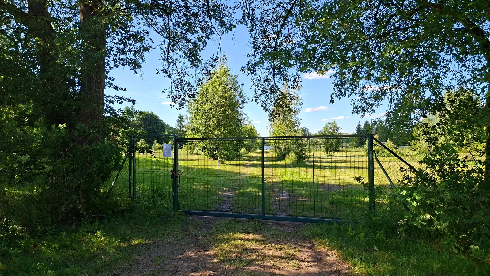 Zdjęcie przedstawia szeroki, poziomy kadr o proporcjach 16:9, ukazujący widok na zieloną, nasłonecznioną polanę zamkniętą metalową bramą ogrodzeniową. Pierwszy plan tworzą gęste korony drzew rzucające cień na ziemię, które z obu stron naturalnie obramowują przejście. Na środku kadru widoczna jest dwuskrzydłowa brama wykonana z zielonej siatki, przez którą widać otwartą przestrzeń z młodymi drzewami i soczystą, zieloną trawą pod błękitnym niebem z rzadkimi chmurami. Na lewym skrzydle bramy przymocowana jest biała kartka z wydrukowanym tekstem, którego treść jest częściowo nieczytelna, ale zawiera nagłówki: „UWAGA”, „TEREN PRYWATNY” oraz „WSTĘP WZBRONIONY”. Całość kompozycji podkreśla sielski, letni krajobraz w pełnym świetle dziennym, a perspektywa prowadzi wzrok w głąb działki poprzez otwartą bramę.